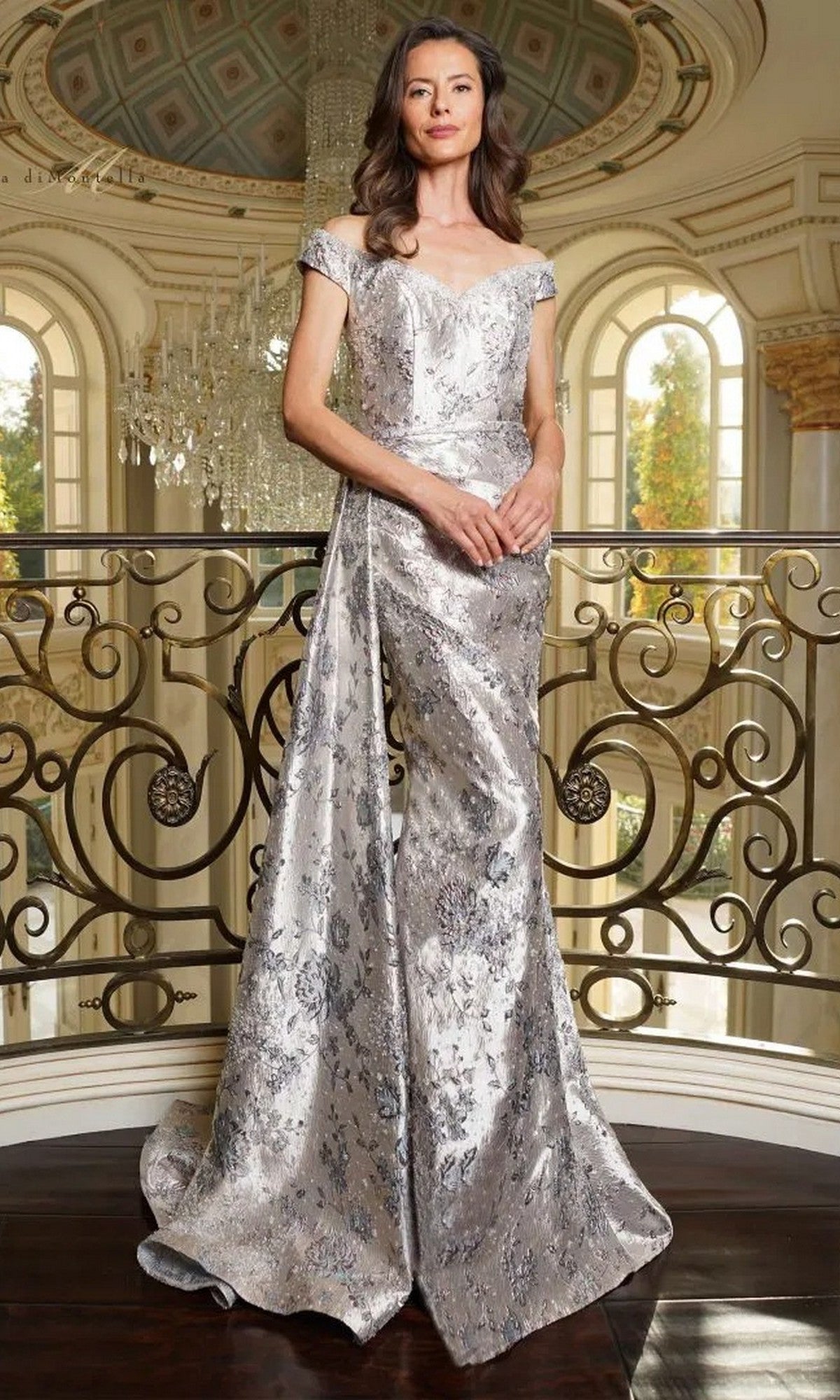 A woman stands indoors in an ornate room with a chandelier and decorative railing, wearing a Rina Di Montella RD2912 long formal dress in blue multi with off-the-shoulder styling and a silver floral jacquard design.
